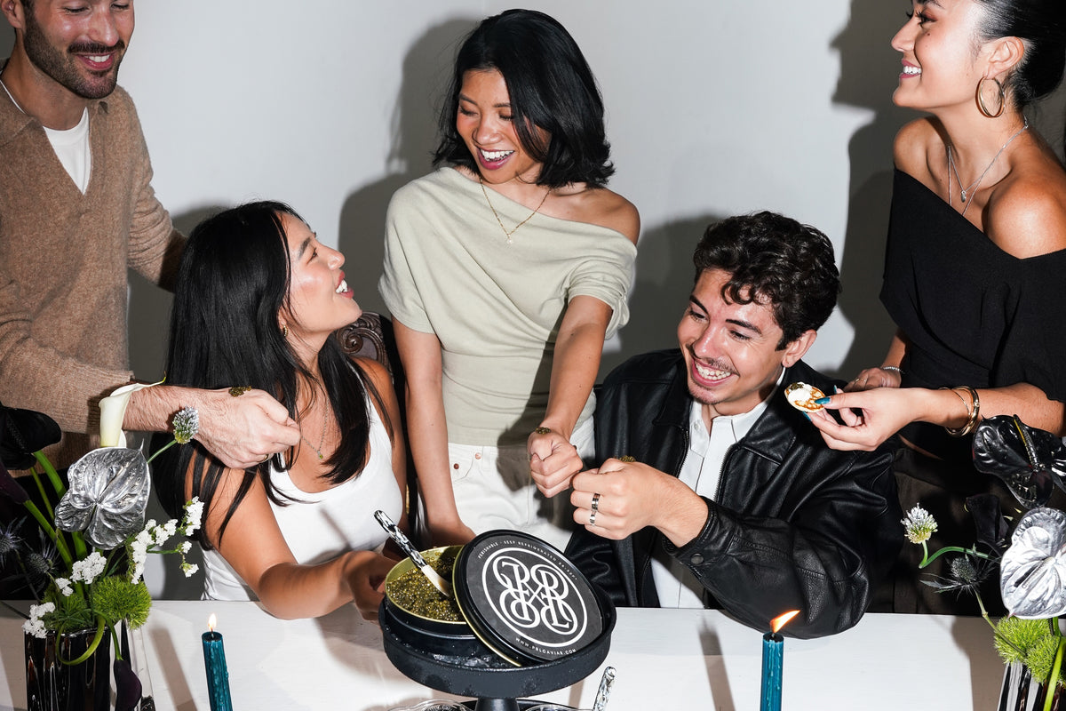 A group of five friends celebrating around a black round cake with a white monogram logo, set against a plain white wall with visible shadows.