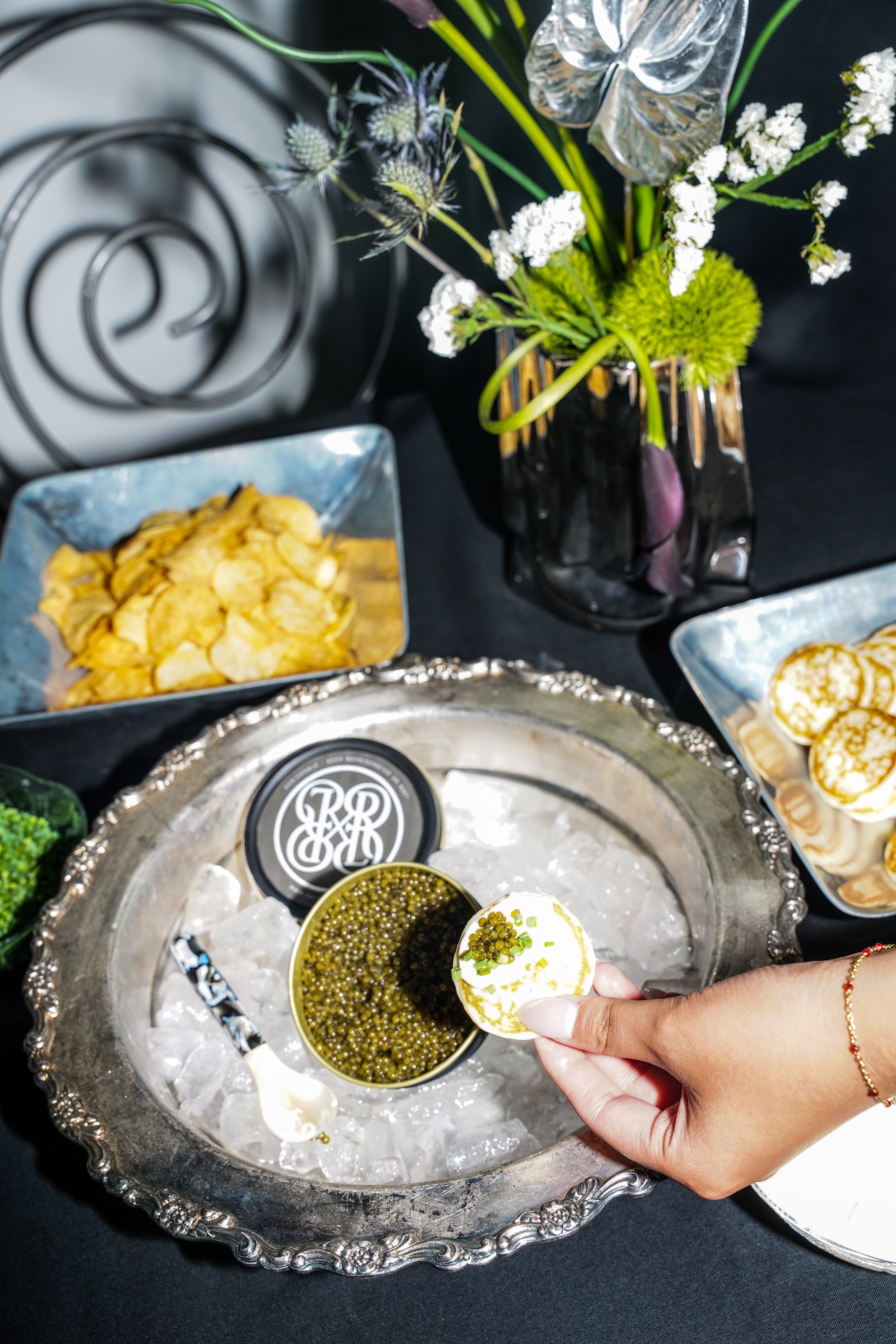 A close-up of a hand holding a small white pastry topped with caviar over a bed of crushed ice on an ornate silver platter, surrounded by chips, pastries, and a floral centerpiece on a dark table.