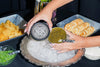 A close-up lifestyle shot of a person's hands holding a black-lidded tin of caviar over a silver platter filled with ice, with a small bowl of green caviar beside it. Surrounding trays include crackers and garnishes on a dark table setting.