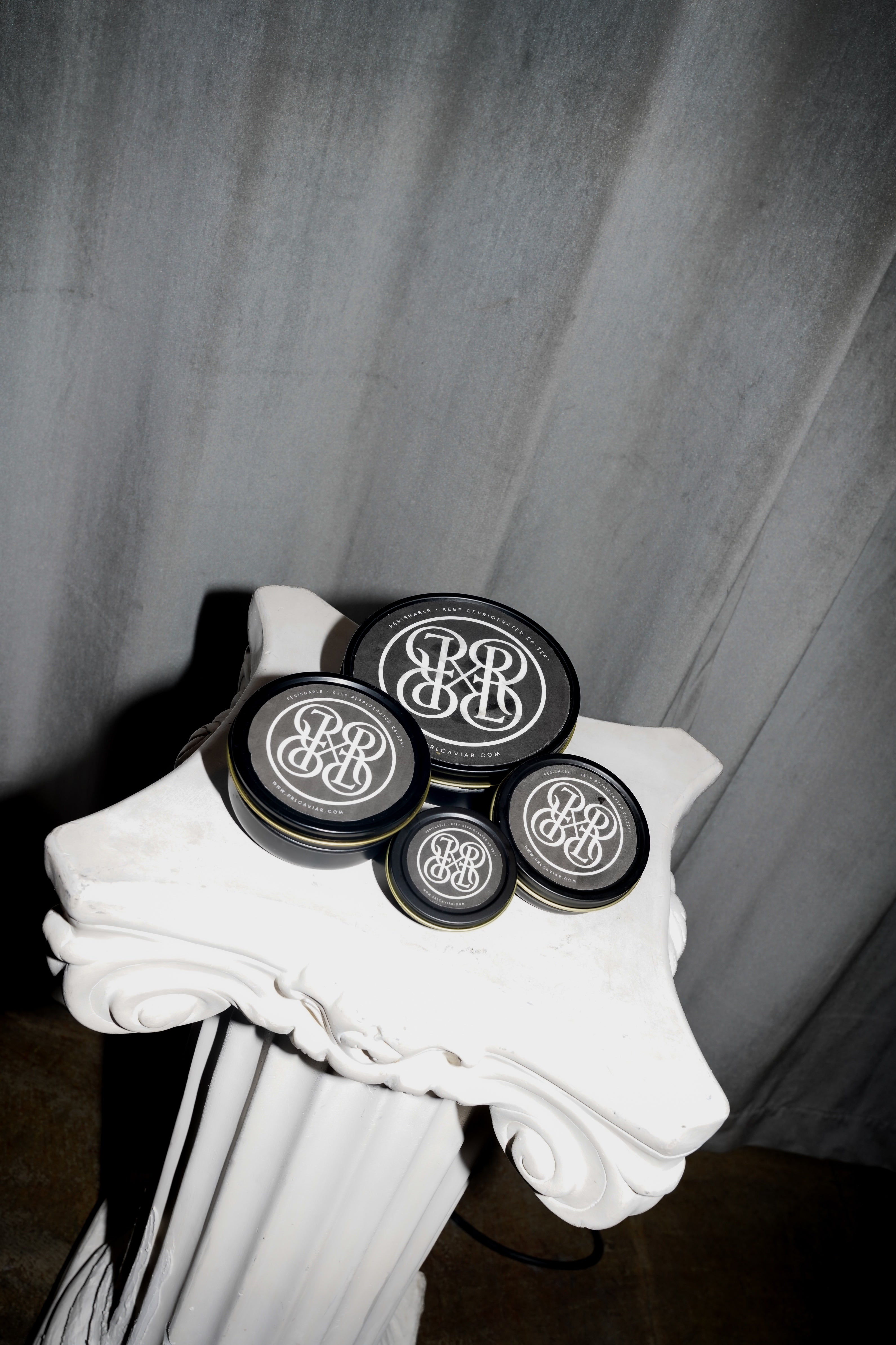 Four round black caviar tins with white monogram logos, arranged on a white decorative pedestal against a gray draped backdrop.