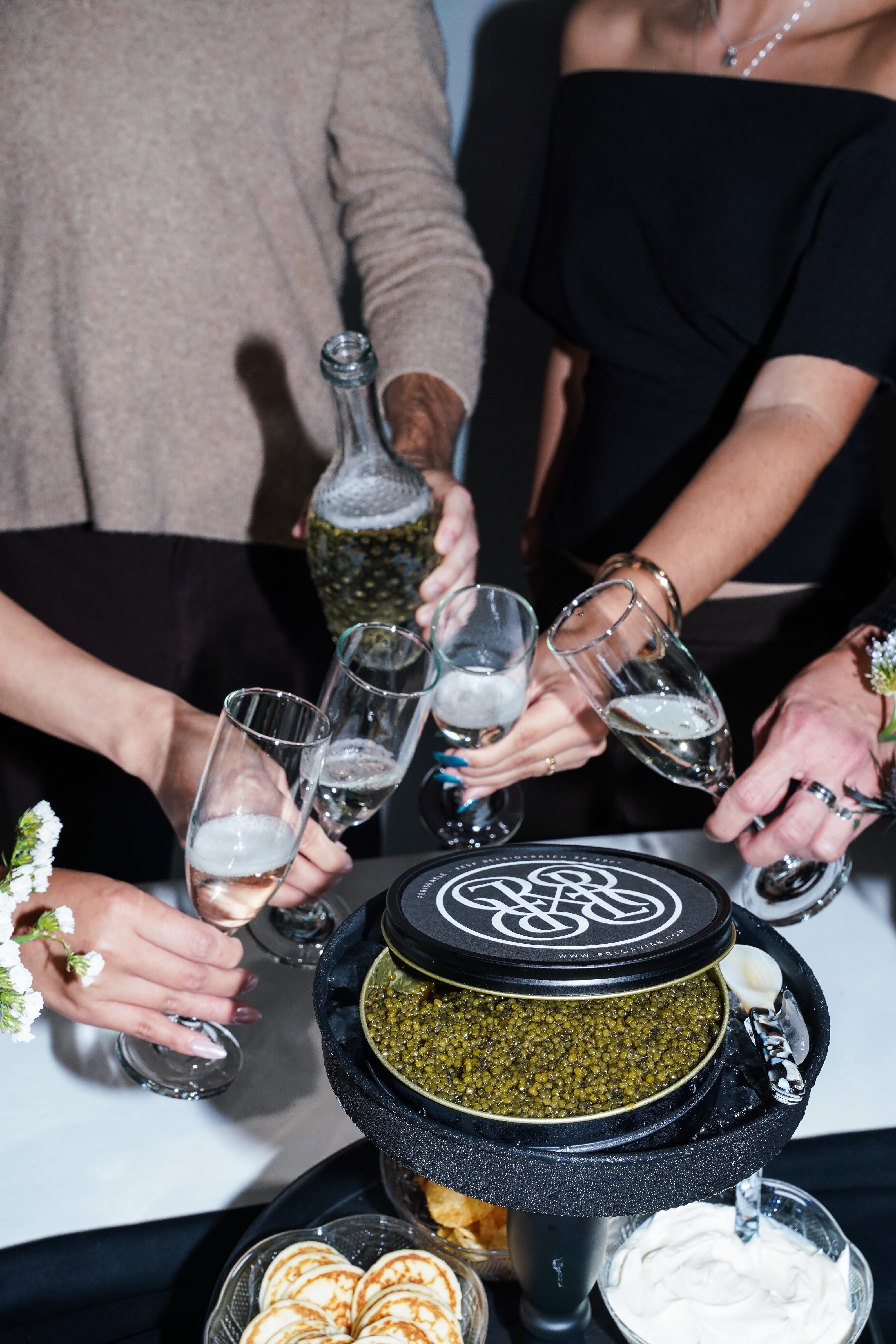 A group of people holding champagne glasses and a bottle around a tiered serving stand filled with caviar, in a social dining setting.
