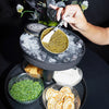 A hand uses a spoon to scoop caviar from a tin resting on a black ice tray, with crackers, a green garnish, and a floral centerpiece on a black background.