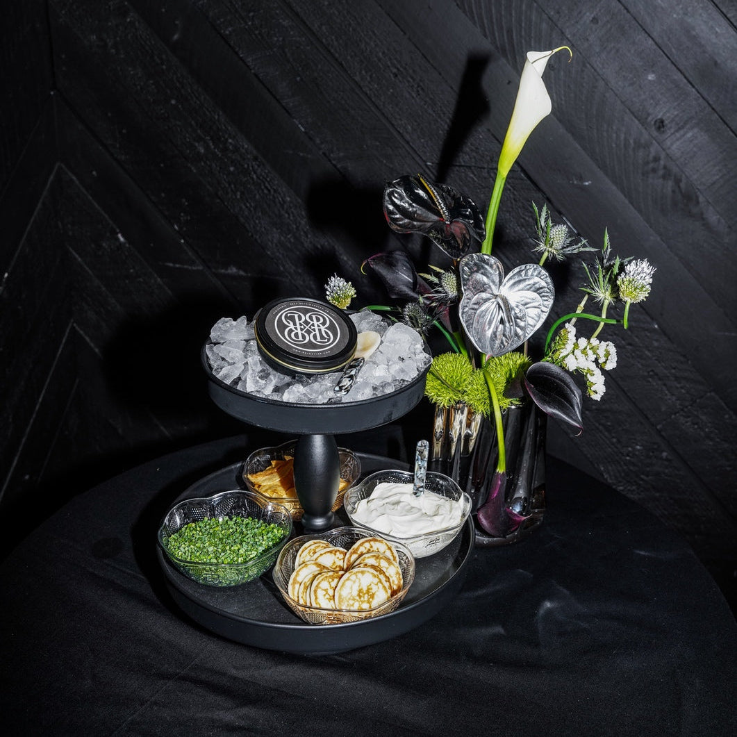A styled black tiered serving tray with caviar and a floral arrangement with a white calla lily, set against a dark wood plank background.