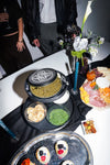 An overhead view of a styled food table with charcuterie, caviar, crackers, and decorated cupcakes on a black tablecloth, with partial people standing around the table.