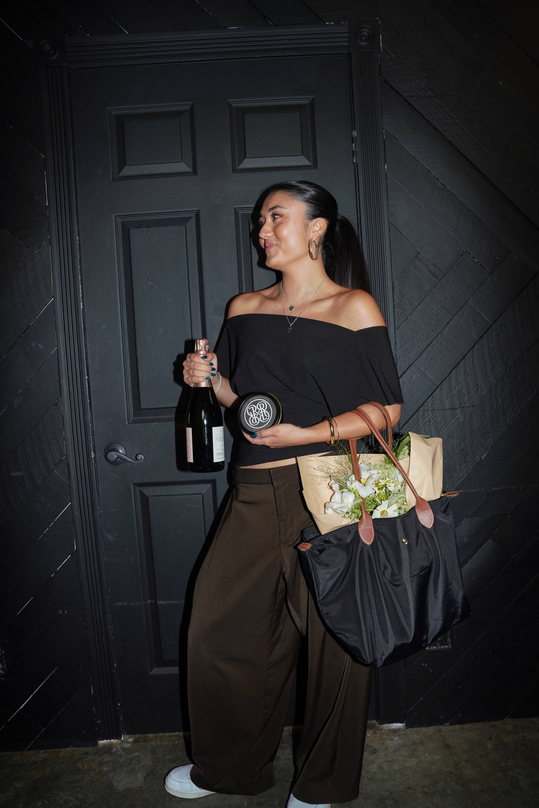 A woman stands in front of a dark paneled door holding a champagne bottle and a small round tin, wearing an off-the-shoulder black top and wide-leg brown pants. She carries a black tote bag and a paper bag with flowers.