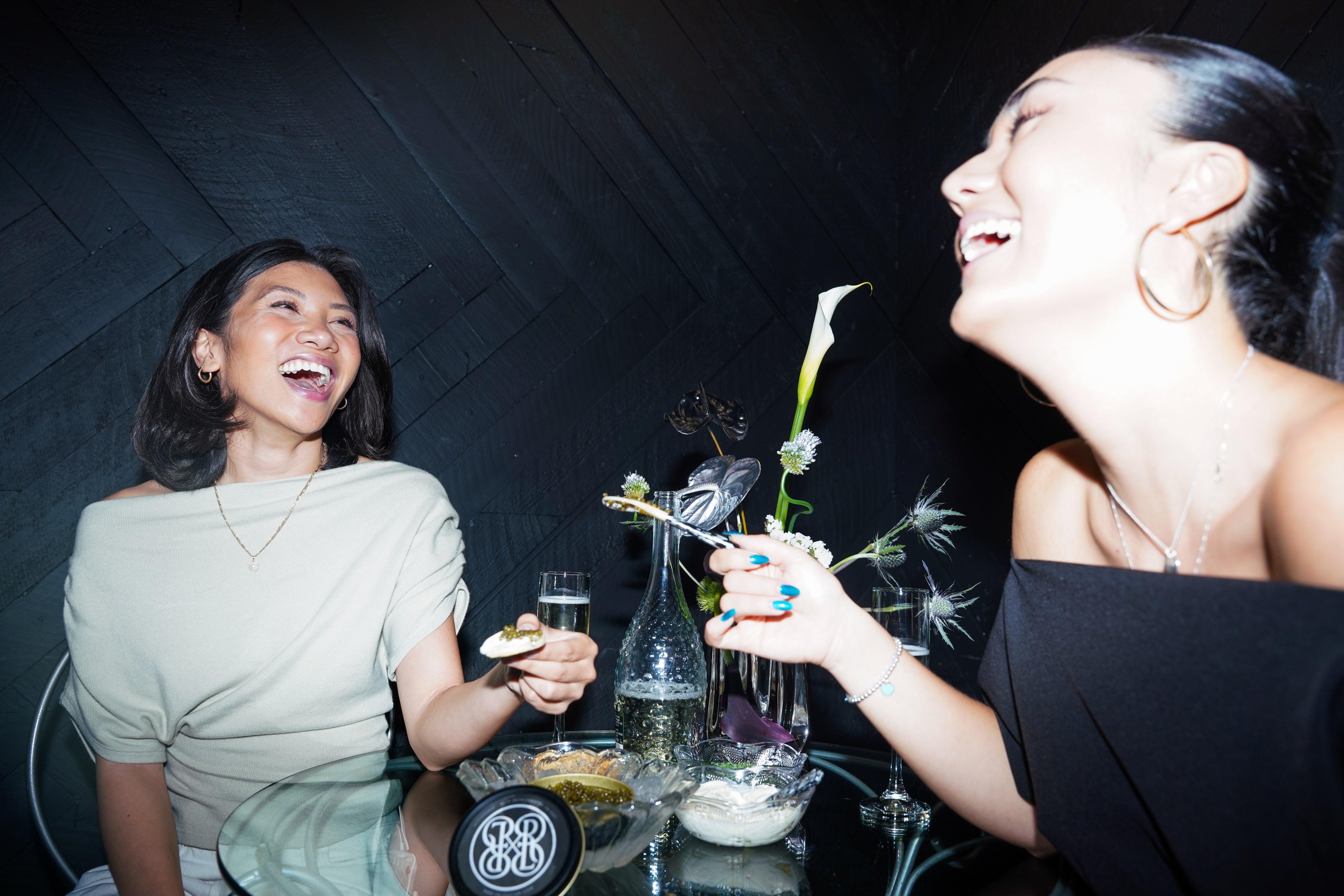 Two women laughing and toasting at a dark table with drinks and flowers in a dimly lit setting.