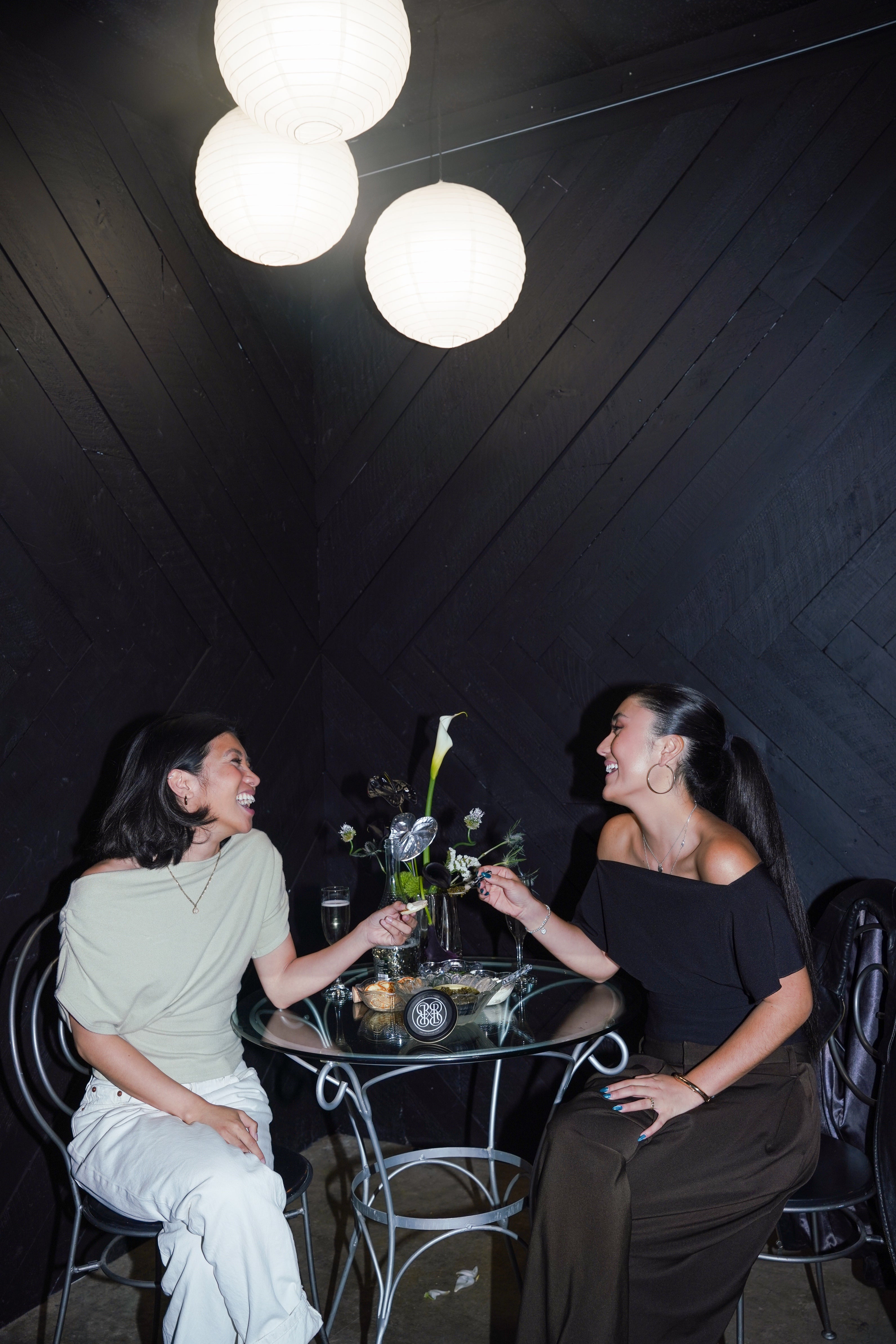 Two women laughing and clinking glasses at a small round table in a dimly lit indoor setting with dark wood walls and hanging white paper lanterns.