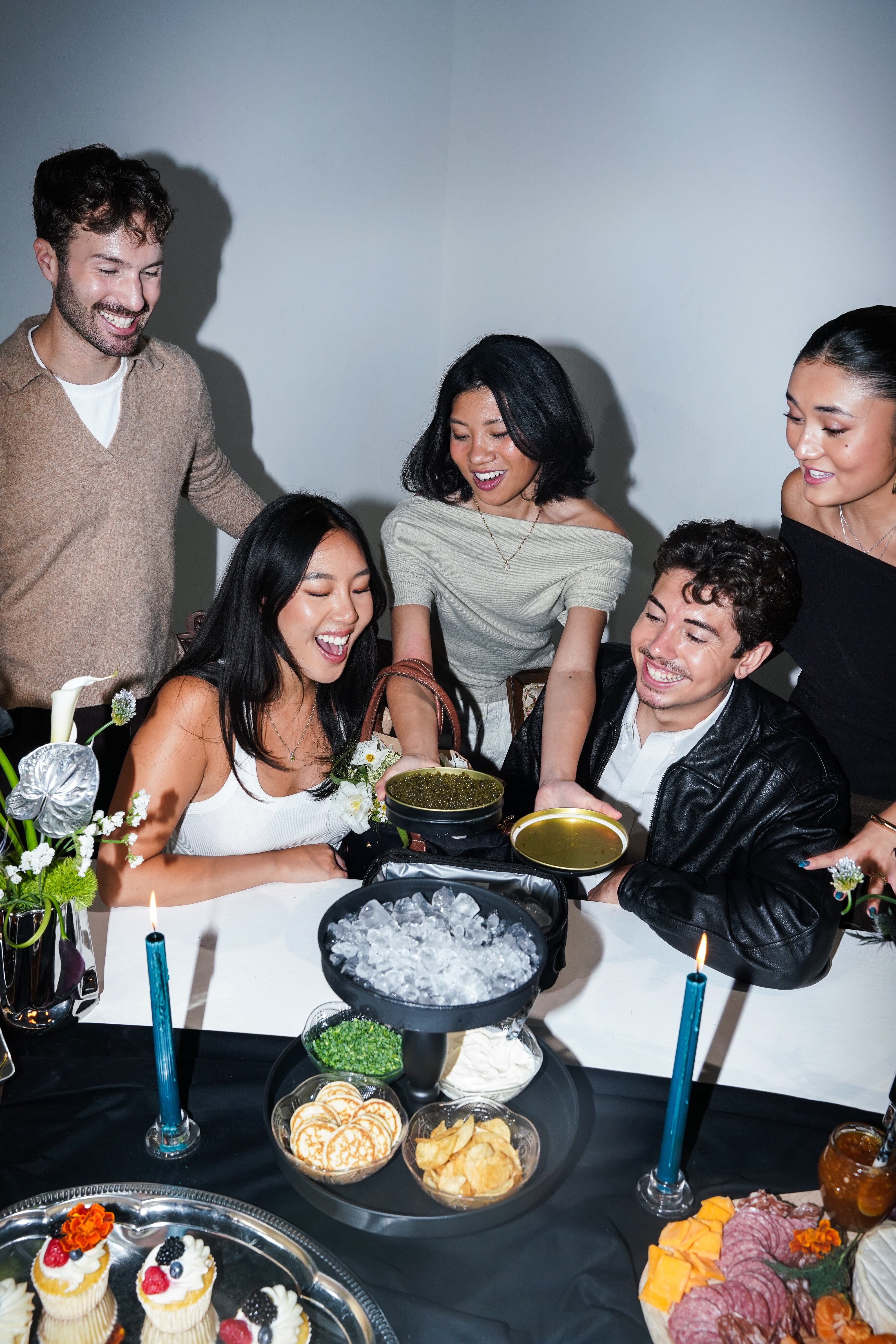 A group of five friends gathered around a table at a party, sharing food and drinks. The central focus is a black tin filled with ice on a tiered stand, surrounded by snacks, candles, and the group laughing and smiling.