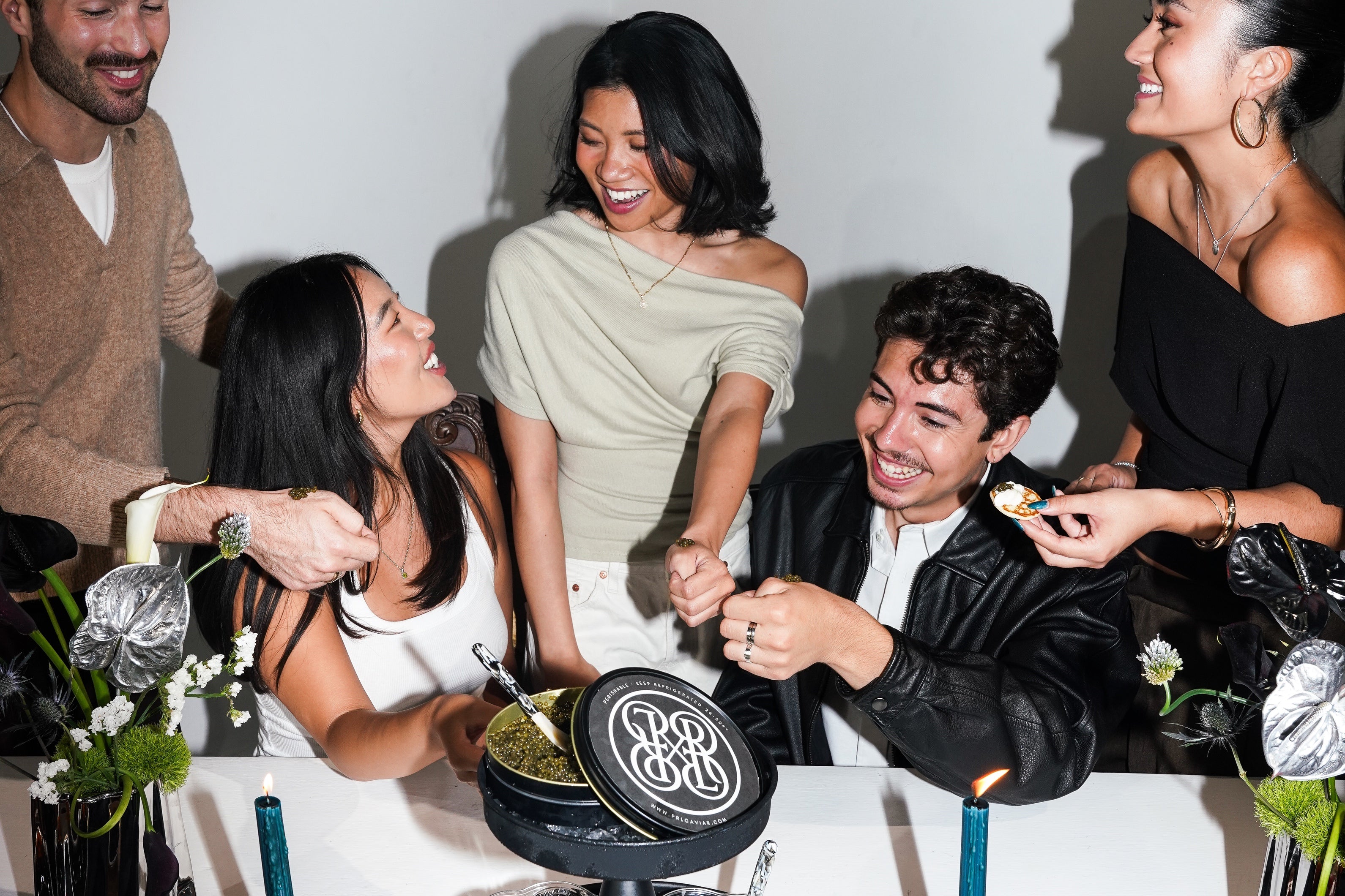 A group of five friends celebrating around a black round cake with a white monogram logo, set against a plain white wall with visible shadows.