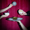 Three hands holding food items against a deep magenta-red curtain background. One hand holds a round tin with a circular logo, another holds a small white dish with a spoon, and a third holds a small bowl of pastries.