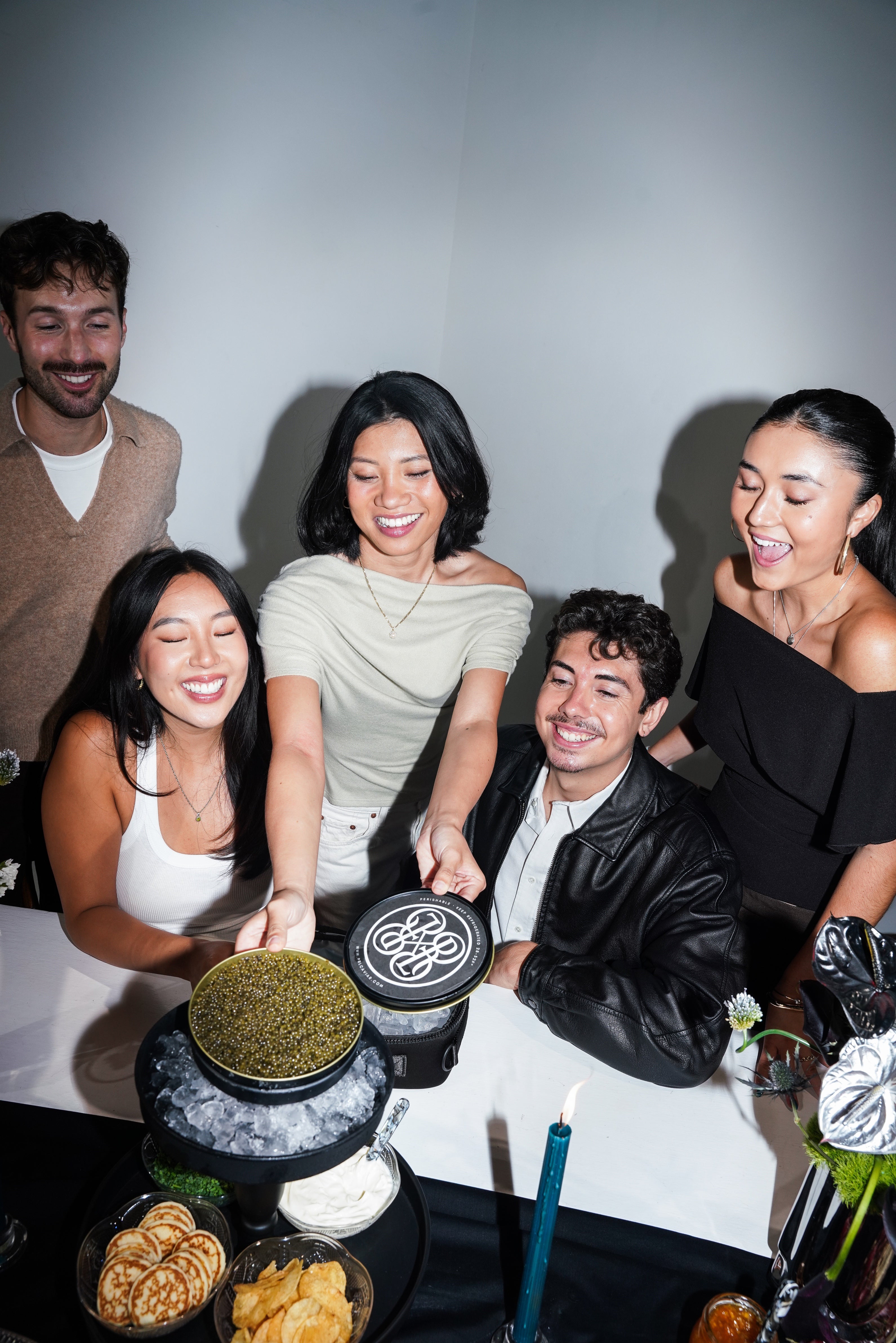 Four people gathered around a table at a dinner or party, smiling and laughing while sharing food and a round black container with a white circular logo on top. The scene is indoors with a plain light wall background and table setting including candles, snacks, and plated food.