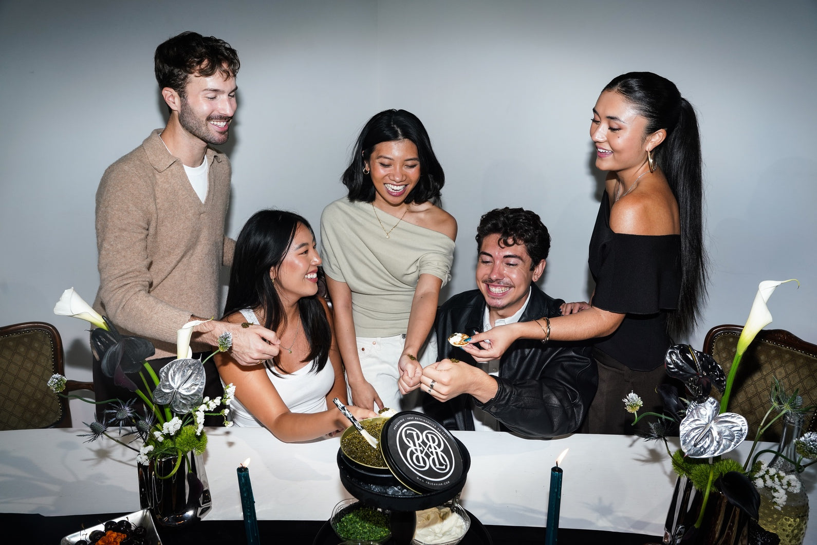 Five people gathered around a table at a dinner or event, smiling and interacting. The setting includes a white tablecloth, floral arrangements, candles, and a black tin with a caviar product.