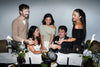 Five people gathered around a table at a dinner or event, smiling and interacting. The setting includes a white tablecloth, floral arrangements, candles, and a black tin with a caviar product.